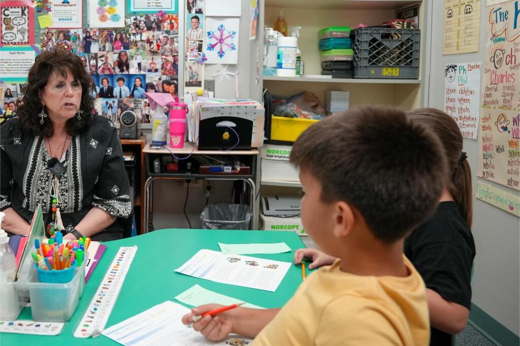 Teacher and two students in a classroom with papers, pencils, and a table with a green tablecloth.