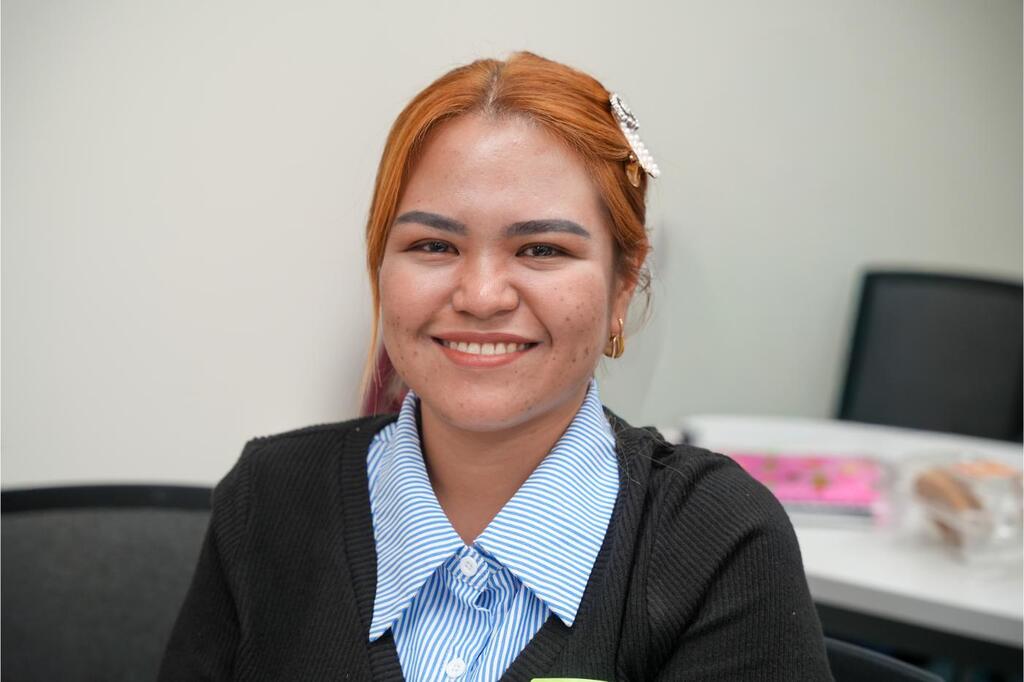 Woman with red hair smiling, seated in an office. She wears a blue and white collared shirt with a black cardigan.