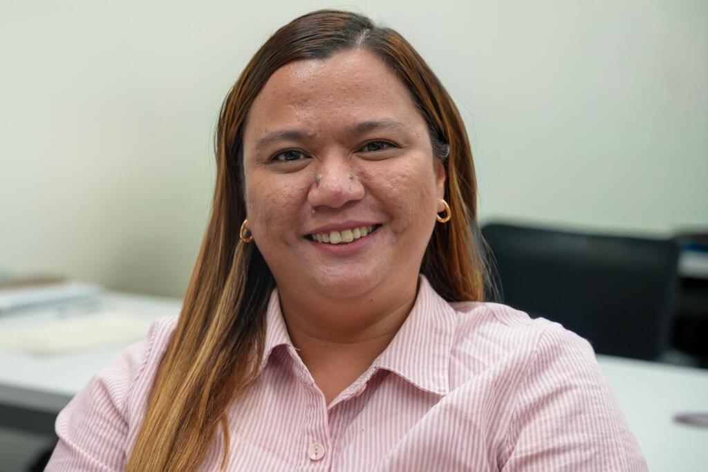 A woman with long hair smiles at the camera. She wears a pink shirt. Behind her, a white desk has a black chair.