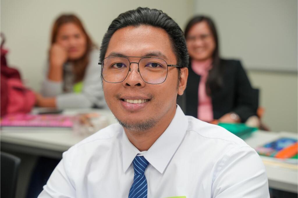 A man in glasses and a tie smiles at the camera, sitting at a desk. Two women, one seated and one standing, are behind him.