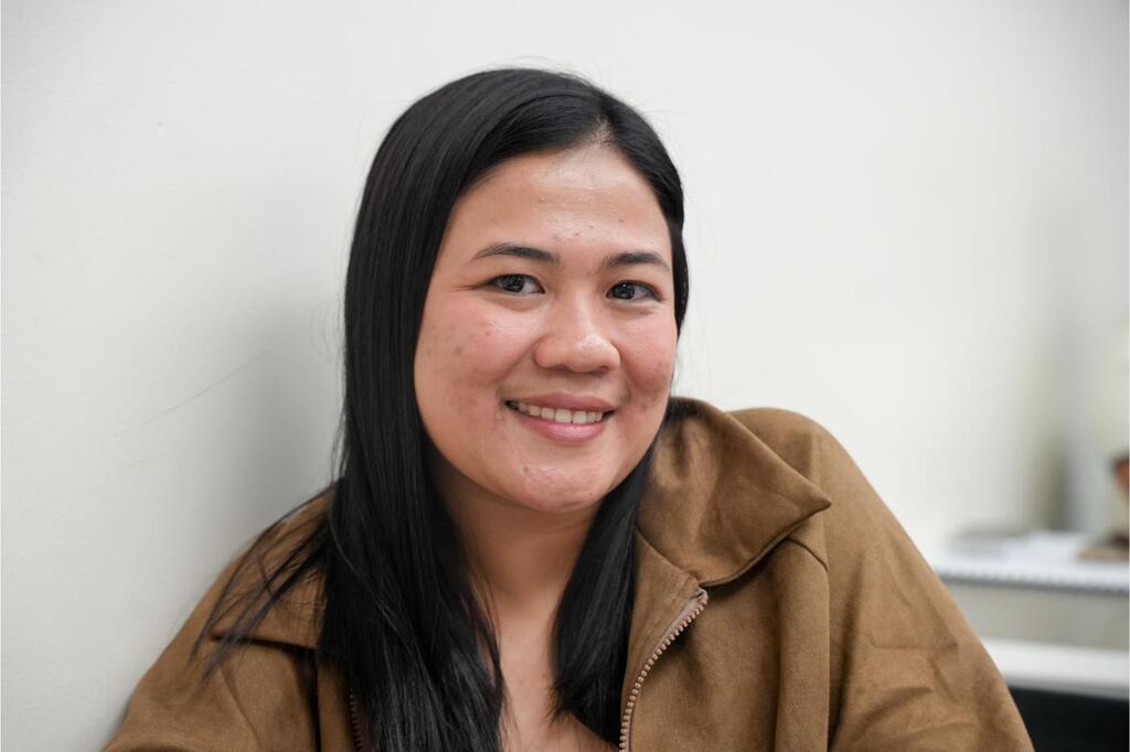 A woman with long dark hair smiles, wearing a brown jacket. Behind her is a plain white wall.