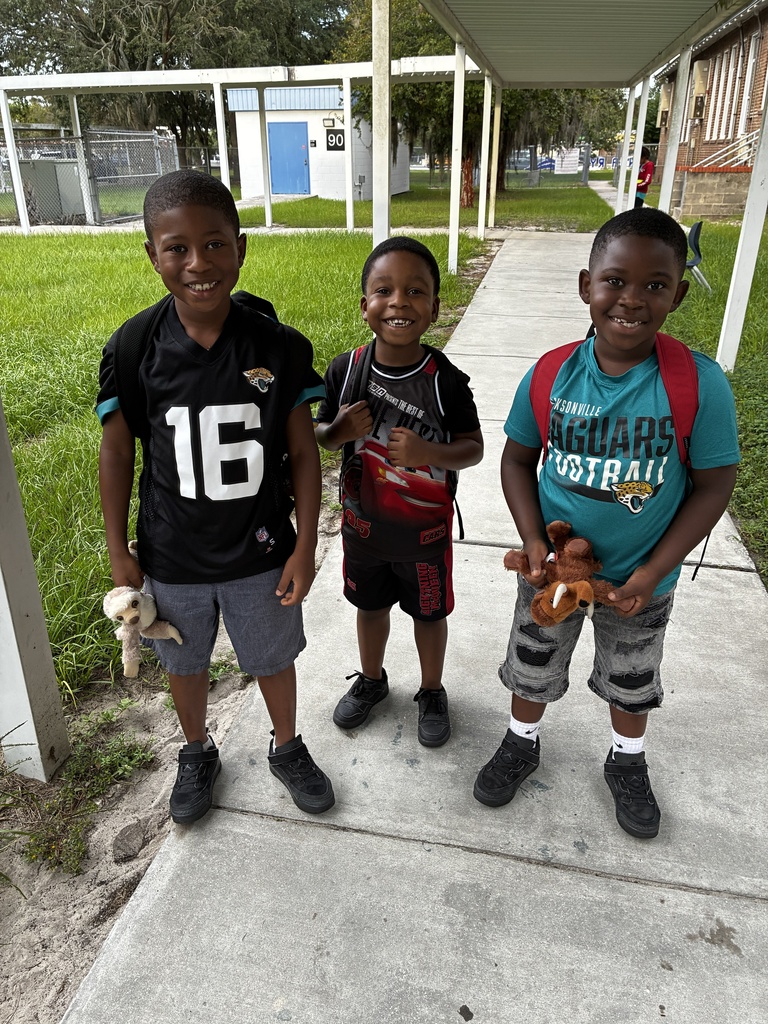 Three children stand on a concrete sidewalk, two holding stuffed animals. One wears a black shirt with "16" and gray shorts, another a red shirt with gray shorts, and the third a teal shirt with red shorts.