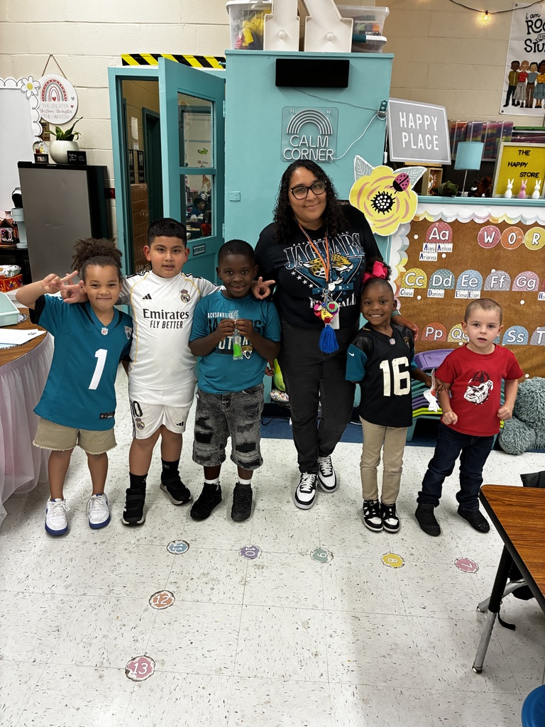 A woman and five children stand together in a classroom with sports jerseys, white floor, and colorful decorations.