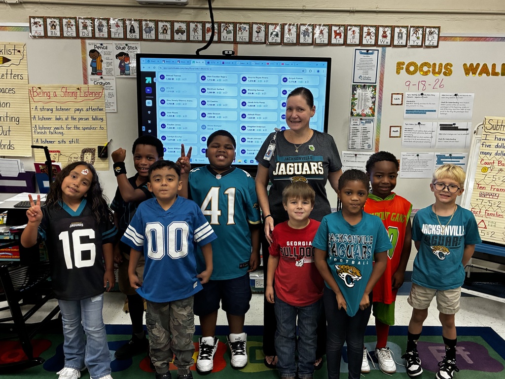 A group of young students wearing sports jerseys stand in front of a classroom projector screen.