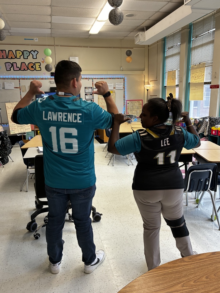 Two adults in a classroom with desks and chairs. A man wears a blue jersey saying "LAWRENCE 16" and a woman in black says "LEE 11".