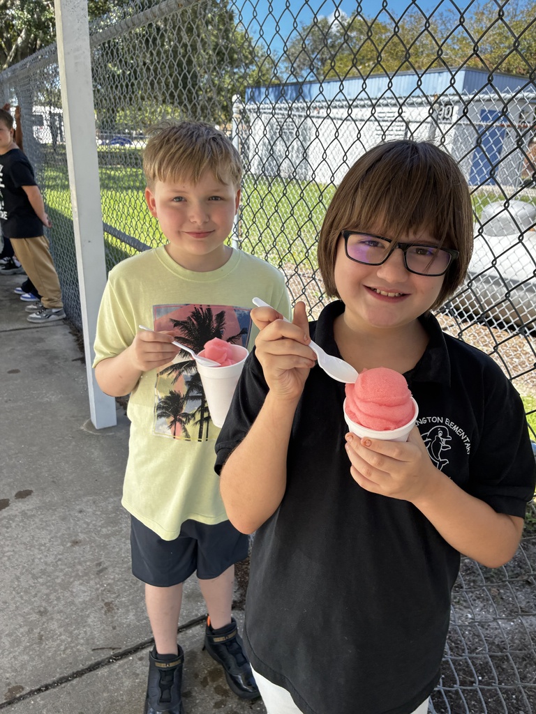 Two children hold ice cream in pink cups. One wears a yellow shirt, and the other wears glasses and a black shirt. A fence and parked cars are in the background.