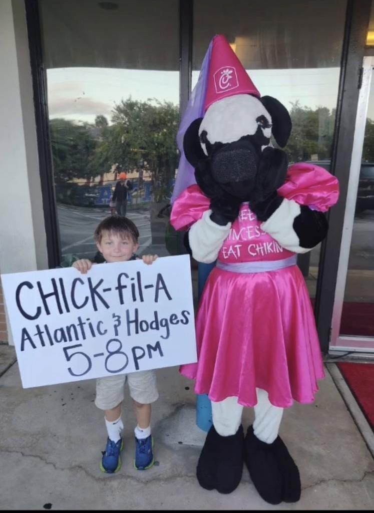 A child holds a sign near a mascot in pink, both standing in front of a school. 