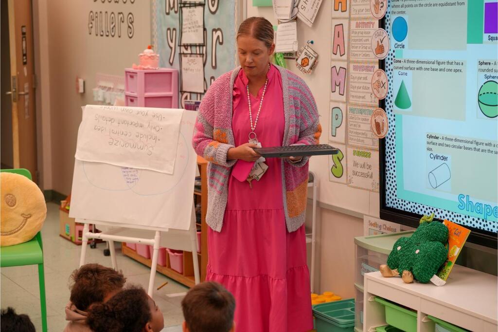 A woman in a pink dress stands in front of a classroom, holding a tablet, teaching children seated around her.