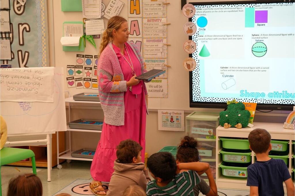 A woman teaching a group of children in a classroom. She stands in front of a projector screen.