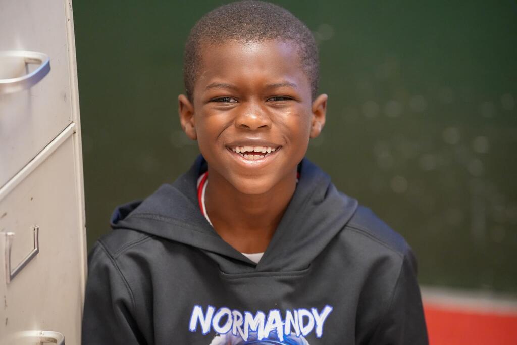 A boy with short hair smiles, wearing a black hoodie with white text. He stands near a metal door.