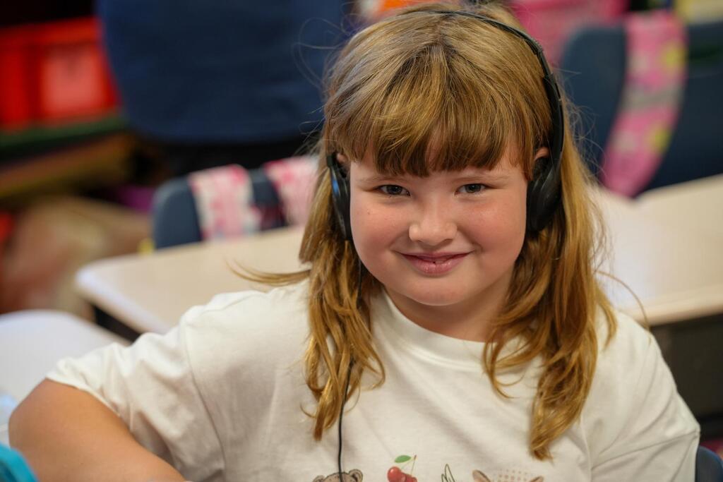 A young girl with long blonde hair, wearing a white t-shirt and headphones, smiles in a classroom.