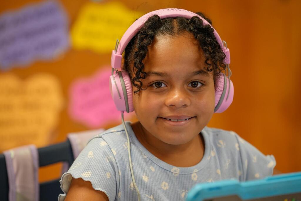 A young girl with curly hair wearing pink headphones smiles at the camera, seated at a desk with a tablet.