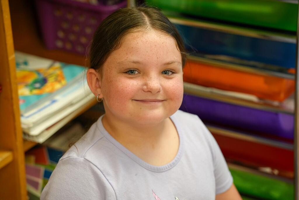 A young girl with a smile poses for a photo. She wears a gray T-shirt and sits in front of colorful folders.