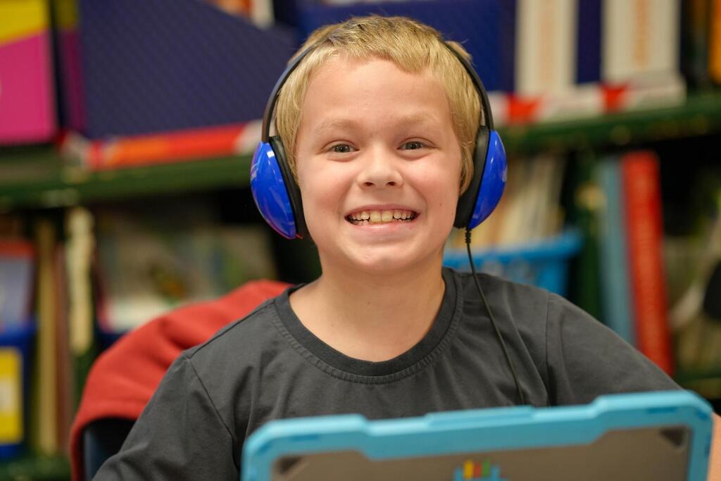 A young boy in a library wearing blue headphones and smiling while looking at a tablet.