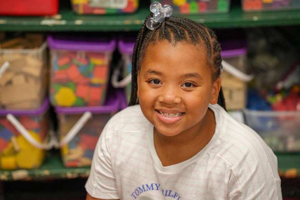 A young girl in a white shirt with Tommy Hilfiger printed on it, smiling. Behind her is a shelf with various colorful storage boxes.