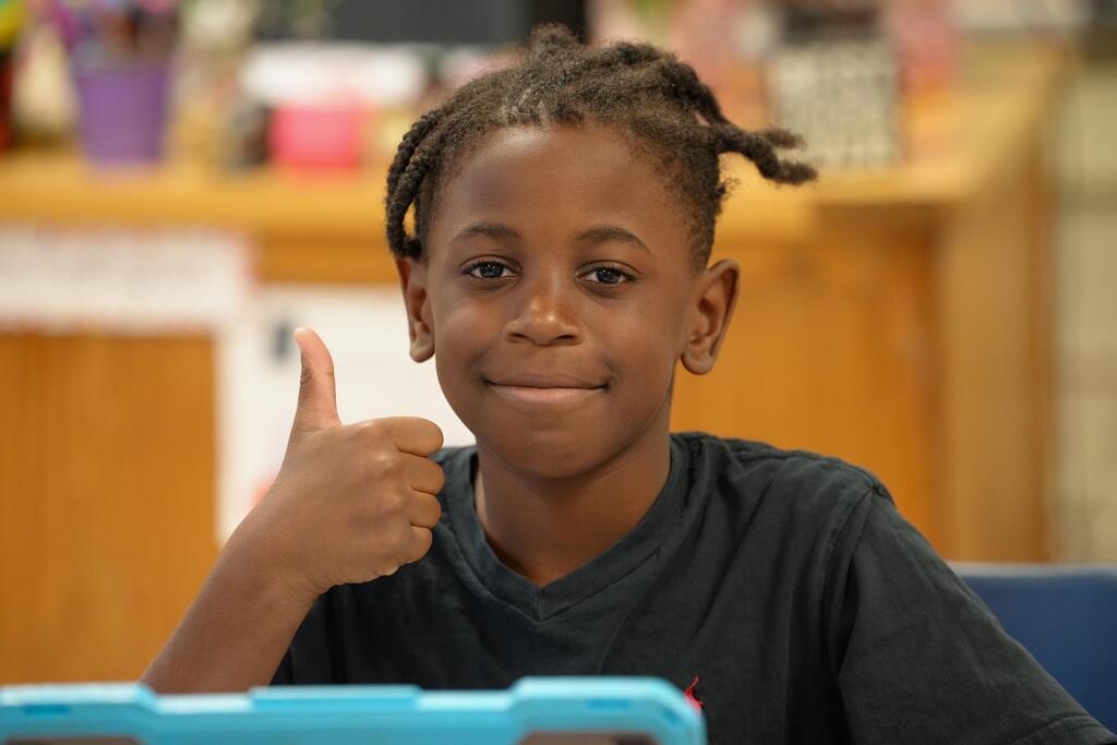 A smiling child sits at a desk, giving a thumbs-up. The desk has a blue tray, and a wooden shelf is in the background.