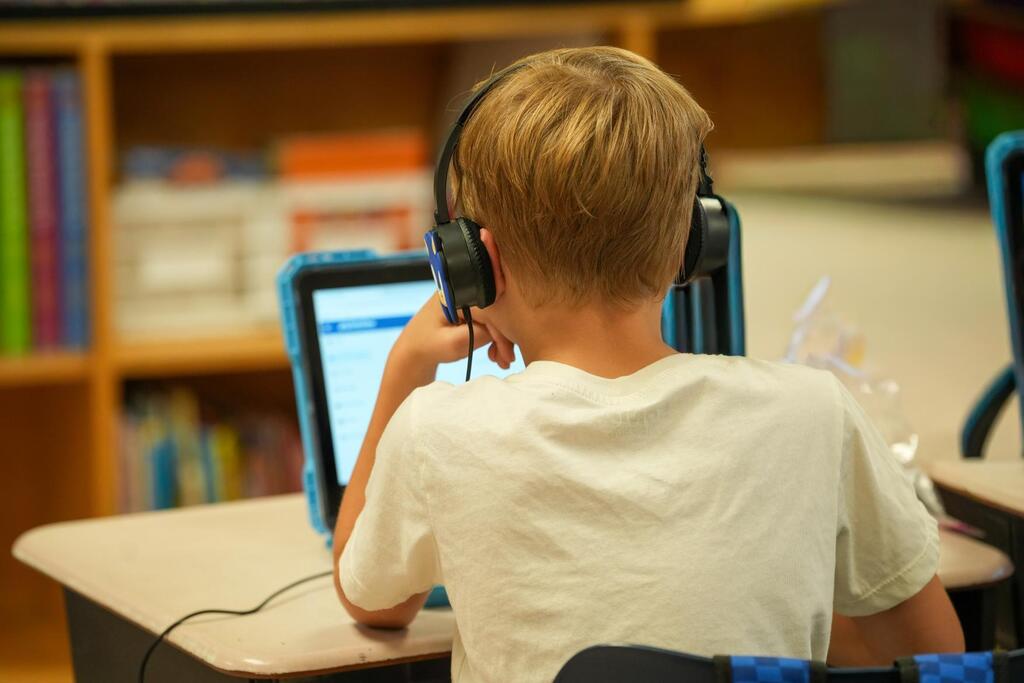 A child wearing headphones uses a tablet at a desk, with a bookshelf in the background.