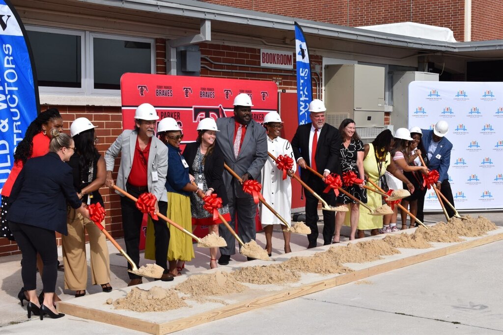 Group wearing white hard hats at a groundbreaking ceremony for a building. Banners on either side.