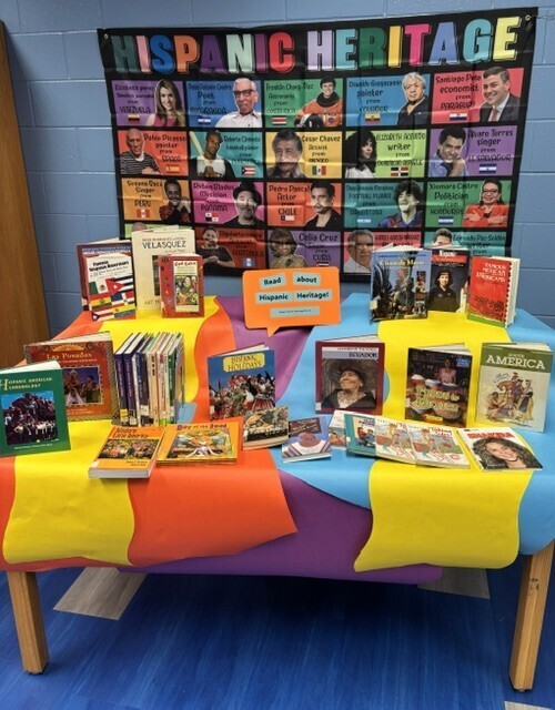 A table covered in a colorful tablecloth, displaying various books. Behind it, a wall features a bulletin board with a "Hispanic Heritage" poster.