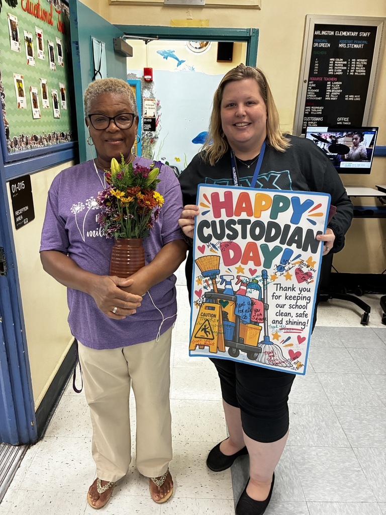 Two women stand in a hallway. One holds flowers, and the other holds a sign that reads "Happy Custodian Day."