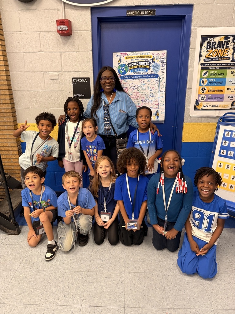 Group of children wearing blue shirts. A woman stands behind them in a hallway. Posters on blue wall.