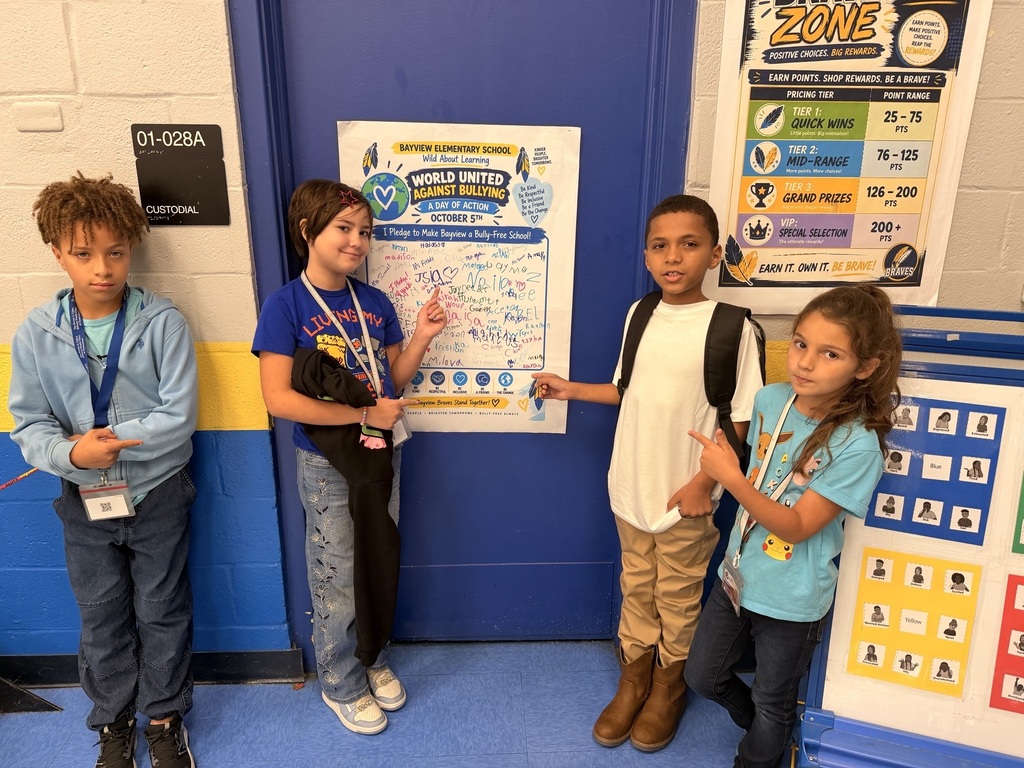 Four children stand in a room with a blue wall. Three hold a whiteboard, and one points at it.