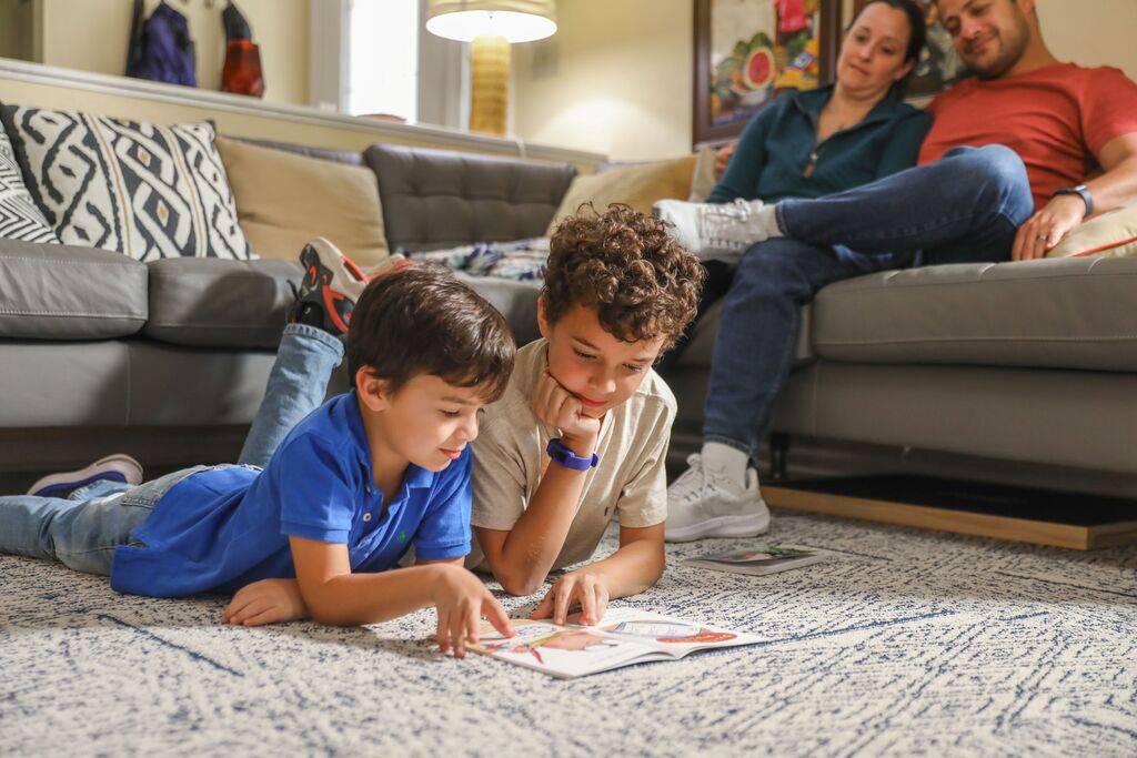 Two young boys read a book together on the carpet in a living room. A man and woman sit on a couch behind them.