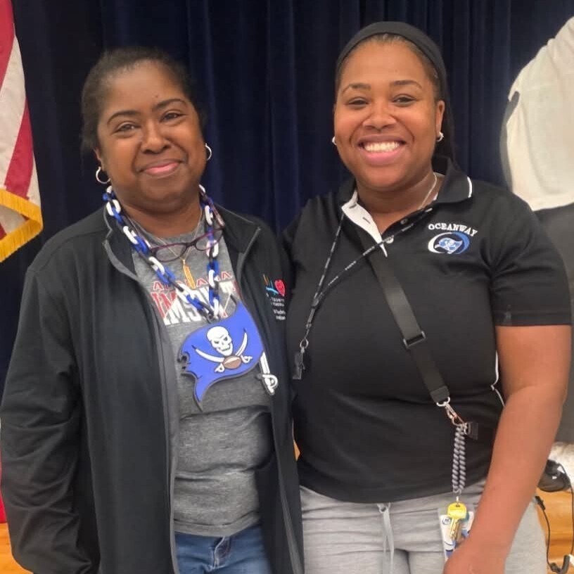 Two women stand together, smiling. One wears a black jacket, the other a black polo with a logo. Behind them is a blue curtain and a flag.