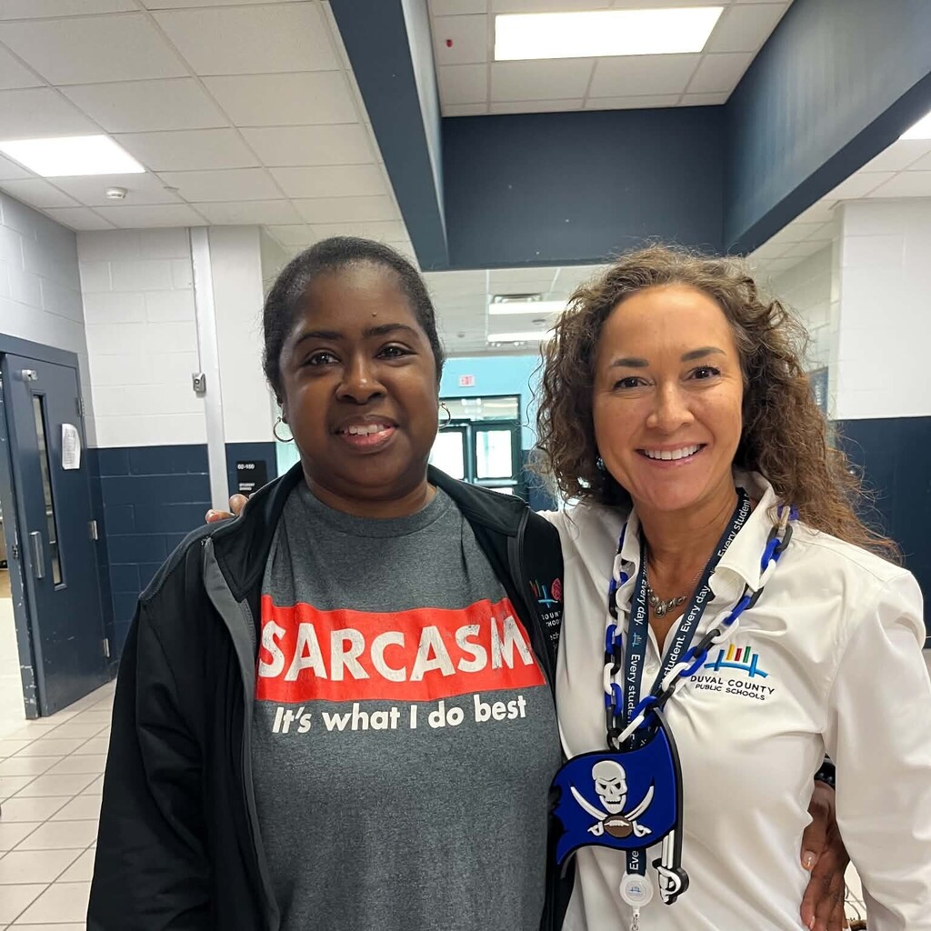 Two women in a hallway, one wearing a gray t-shirt with "SARCASIM" and the other in a white lab coat.