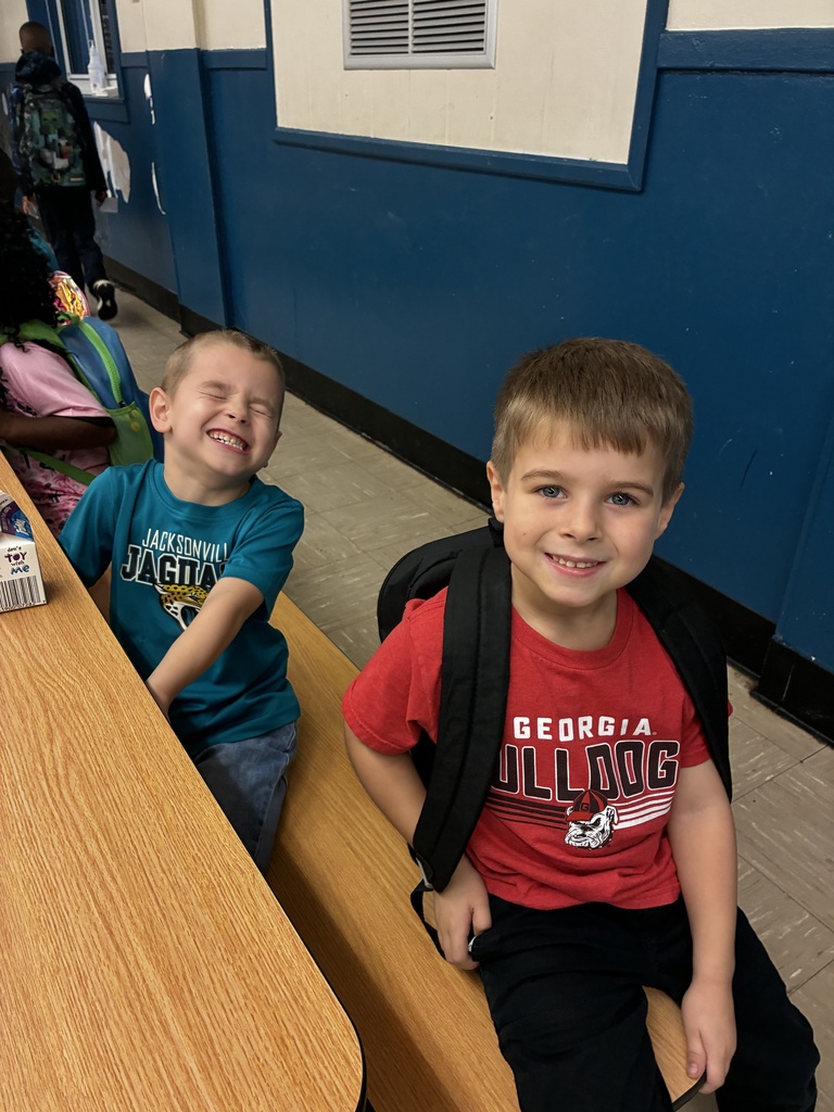 Two boys sit on a bench, smiling at the camera. One wears a blue shirt, the other a red one. Behind them is a blue wall with a person walking.