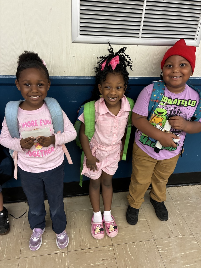 Three kids stand close together. One wears a pink shirt and holds a lunchbox. Another wears a blue shirt and holds a toy. The third wears a red hat and a backpack.