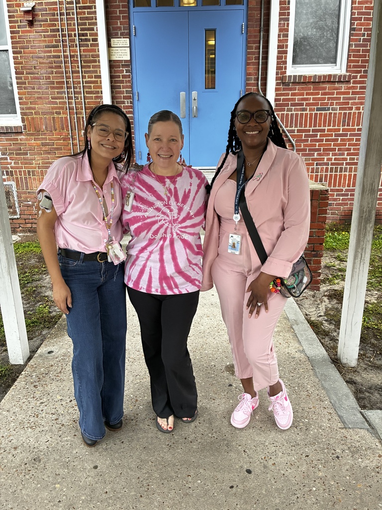 Three women stand together in front of a brick building. One wears glasses and a tie-dye shirt, another wears a pink suit and glasses, and the third wears a pink outfit.