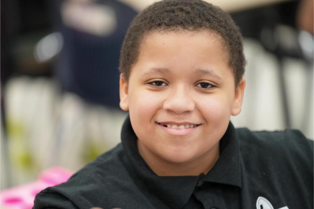 A young person with a smile wears a black polo shirt. Blurry chairs and tables in the background.