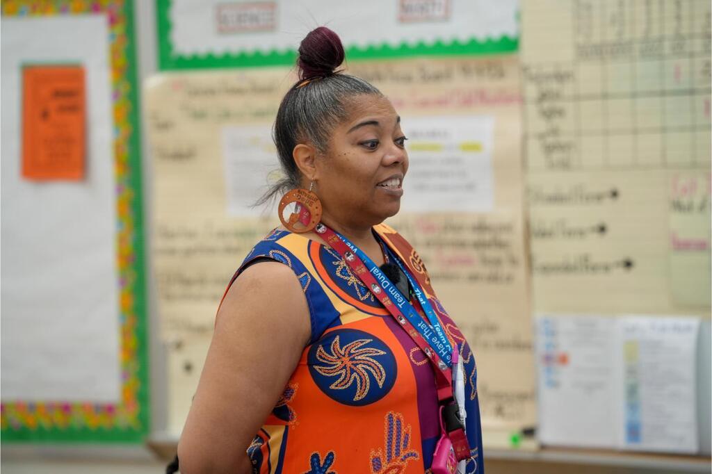 A woman with a bun hairstyle stands in front of bulletin boards, wearing a sleeveless shirt with a graphic design.