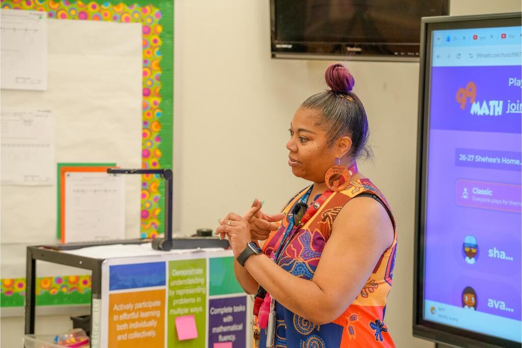Person standing in a classroom, gesturing while speaking. Screen behind them shows 'MATH'. Background includes a whiteboard and projector.