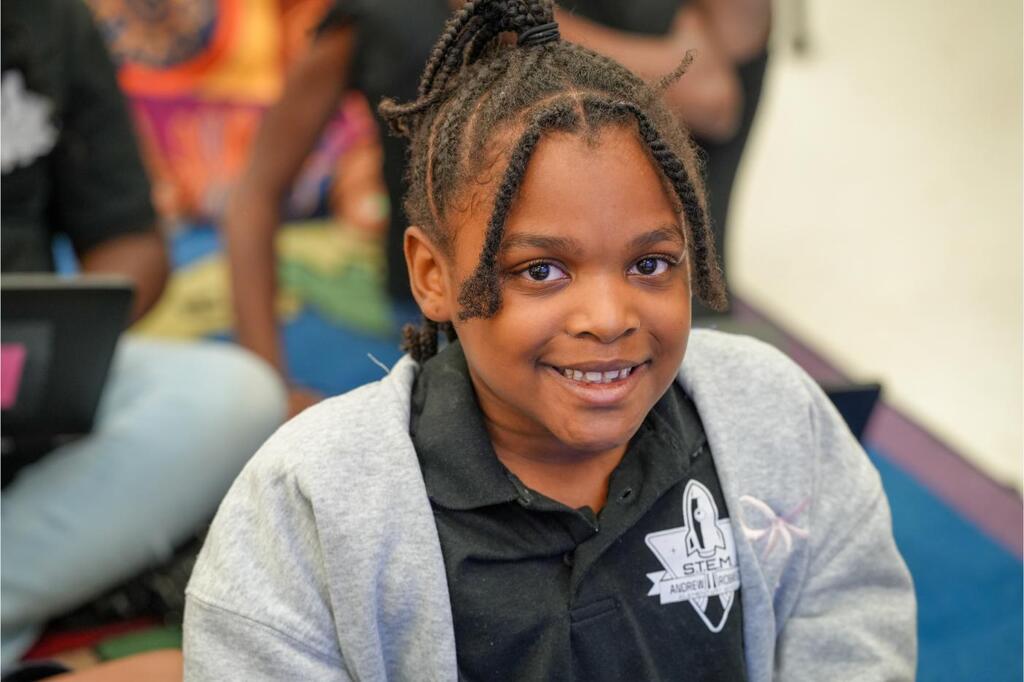 A young girl with braided hair in a black shirt sits in a classroom, smiling. Background includes people.