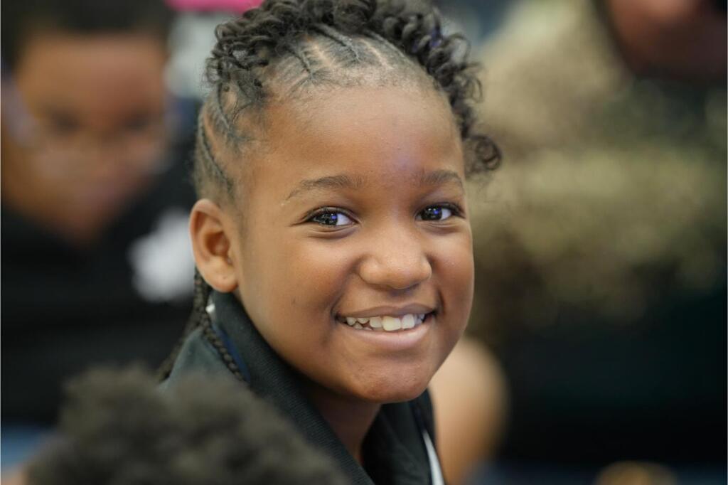 Close-up of a smiling girl with curly hair. She wears a black jacket. Blurred faces in the background.