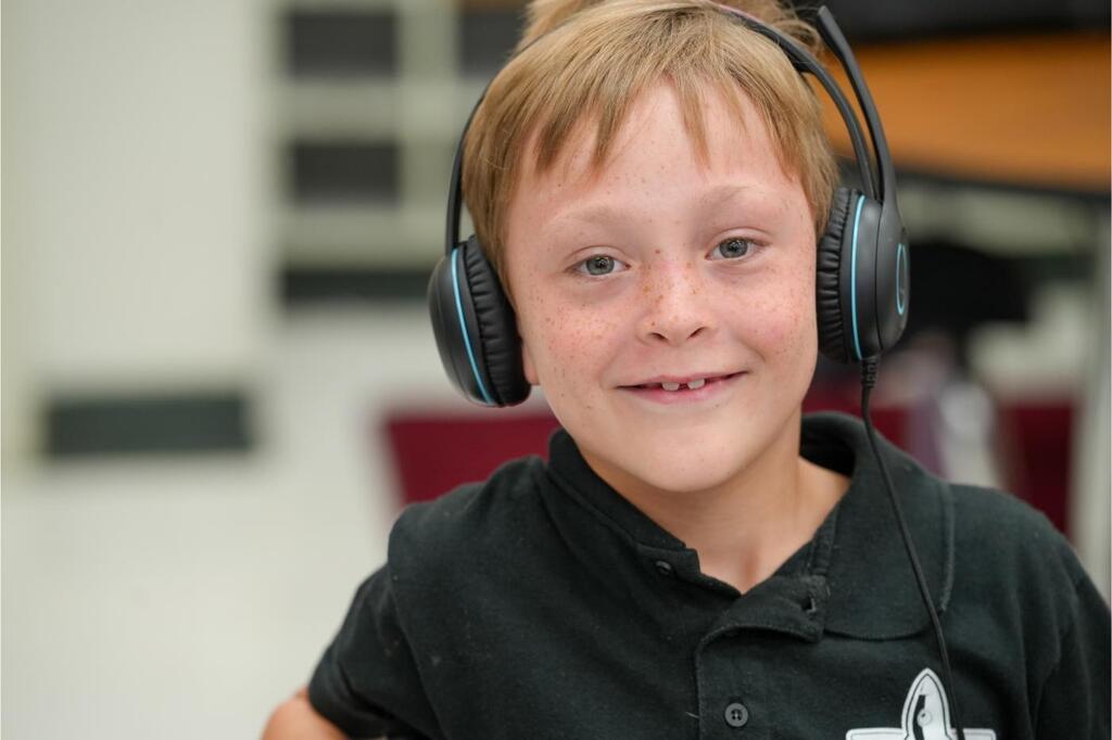 A boy wearing headphones smiles at the camera. He wears a black shirt with a logo on the left chest.