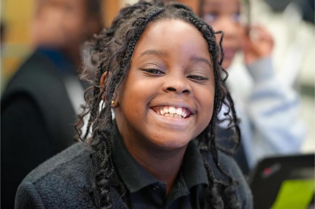 A smiling child with curly hair wearing a black shirt, seated among other students in a classroom.