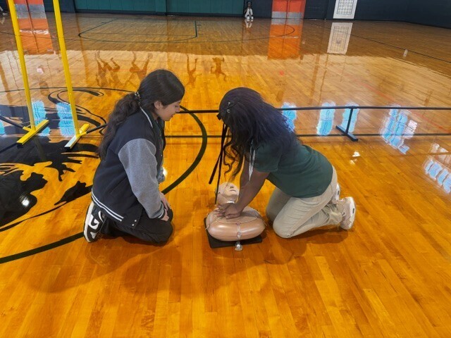 Two individuals on a basketball court, one practicing CPR on a dummy while the other watches.