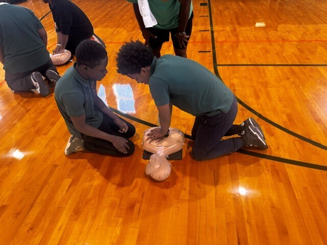 Two individuals are practicing CPR on a dummy on a basketball court. Others are nearby.