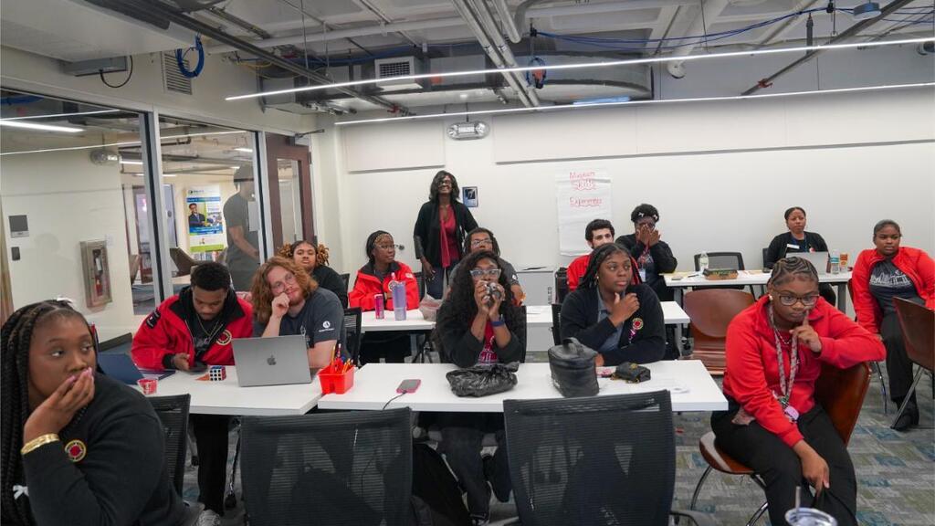 A group of individuals seated at desks, some wearing red shirts, in a classroom setting.