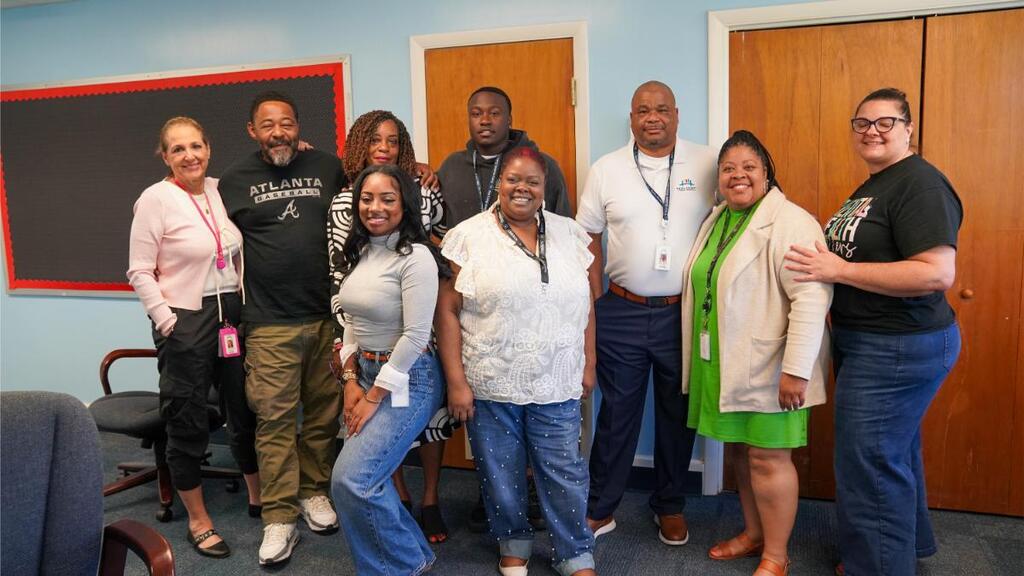 Nine people in casual clothing stand in a room with blue walls, wooden doors, and a chalkboard.