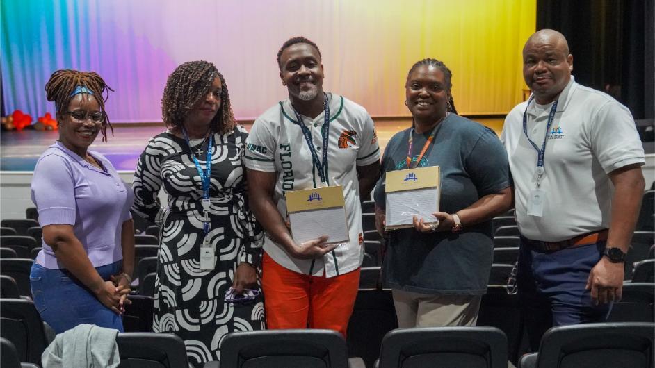Five people stand together holding certificates. One man wears a white shirt and orange pants. The background includes colorful stage curtains.
