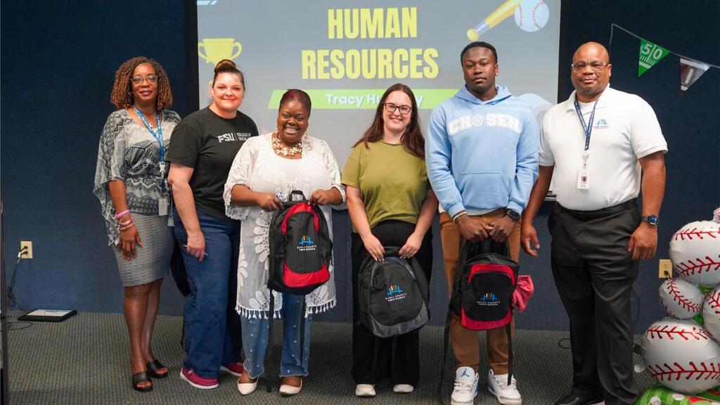 A group of seven people holding backpacks stands in front of a screen with "Human Resources".