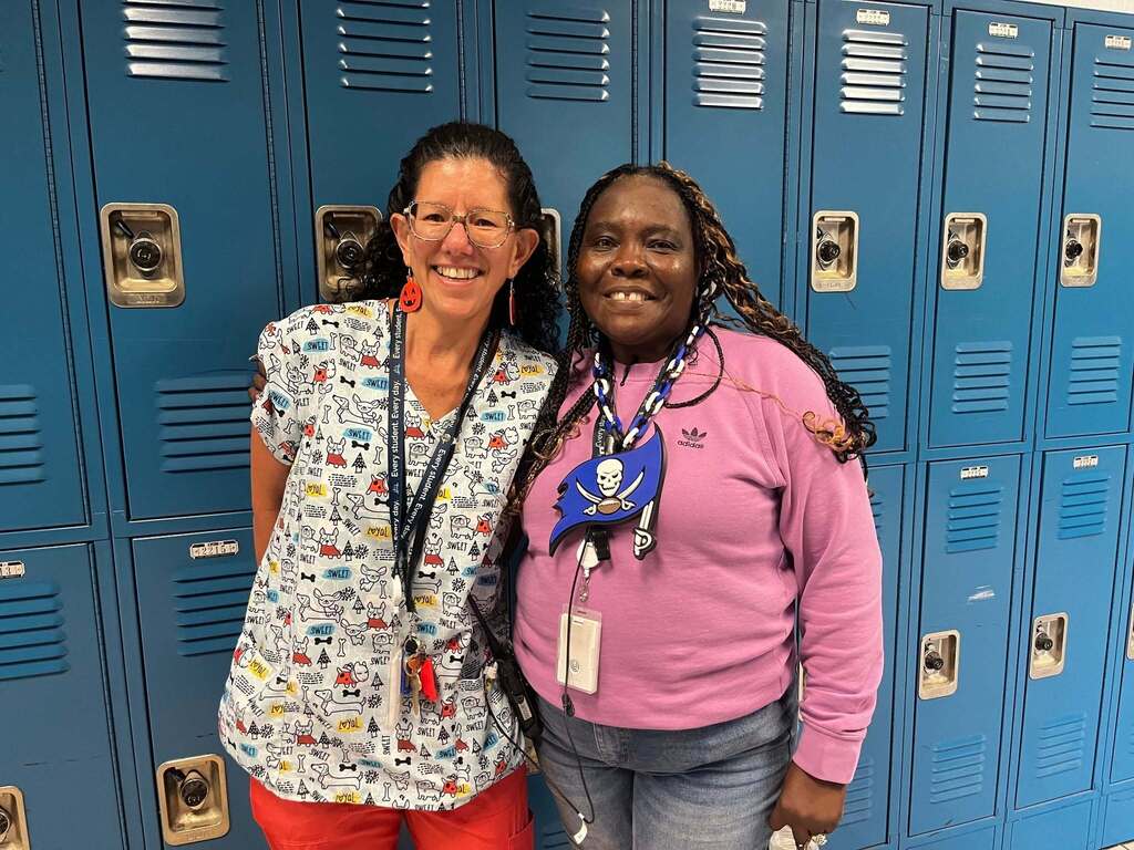 Two women stand in front of blue lockers, smiling. One wears glasses and a printed shirt; the other wears a pink sweater and has a scarf around her neck.