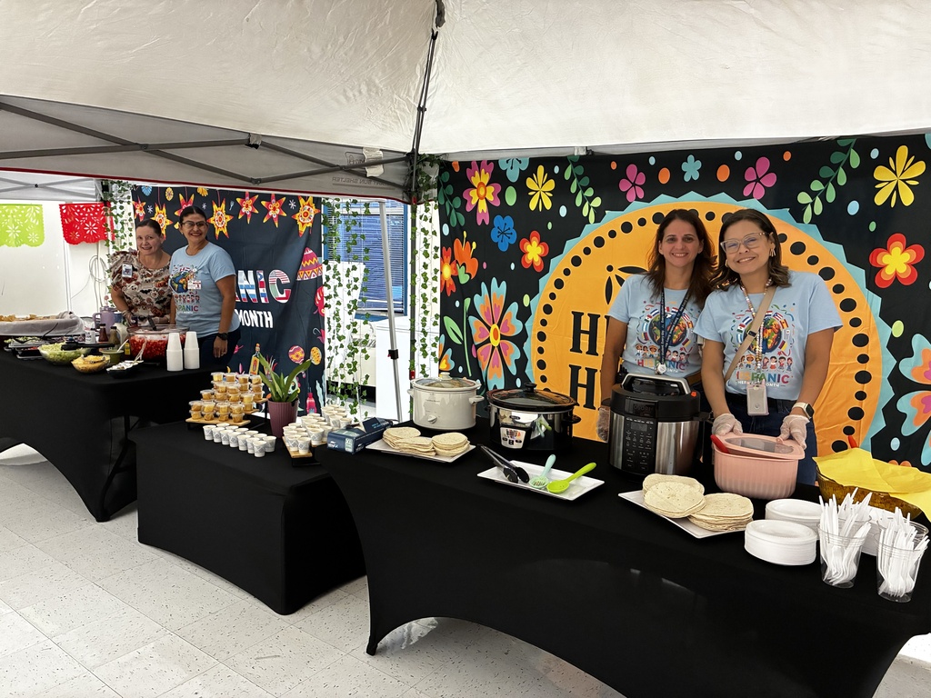 Three women stand behind a table with various foods. Colorful decorations hang in the background.