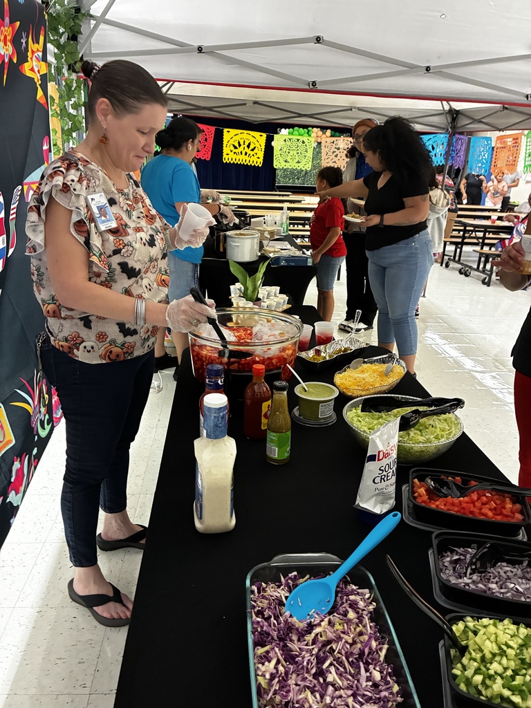 A person prepares food at a table with various salad bowls under a tent with Mexican decorations.