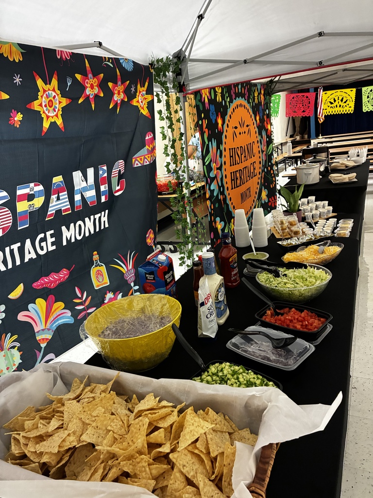 Food display with chips, salsa, guacamole, and other dips. Tent with Spanish Heritage Month banner.