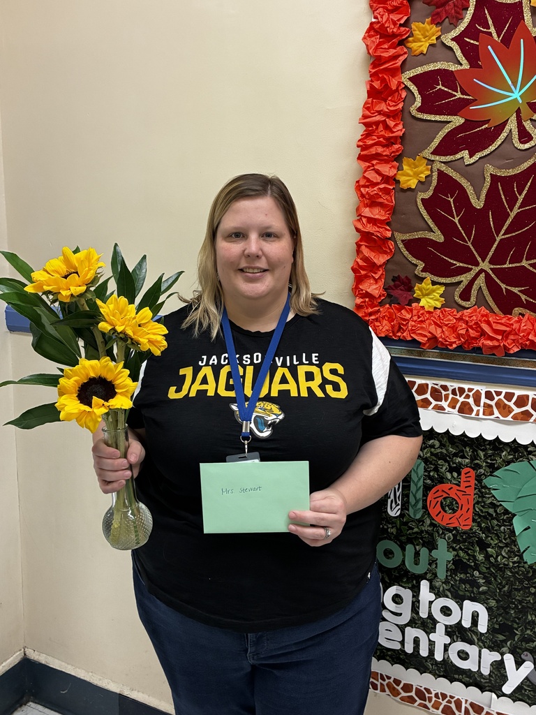 Woman holding a vase with sunflowers and a green card, standing in front of a bulletin board.